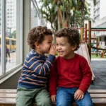 Fotografia em plano médio de dois meninos gêmeos idênticos de aproximadamente 2 anos, sentados em um banco de madeira em uma praça urbana brasileira. O menino à esquerda veste um moletom listrado azul e branco e cochicha algo no ouvido do irmão, que veste um moletom vermelho e ri alegremente. Ao fundo, vê-se o movimento desfocado de uma rua com um ônibus amarelo e árvores, criando um contraste entre a intimidade dos irmãos e o caos da cidade. A imagem ilustra o fenômeno da criptofasia e a conexão profunda entre gêmeos