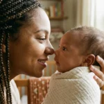 Close-up fotográfico e intimista focado nos rostos de uma mãe e seu bebê recém-nascido, muito próximos e alinhados. A mãe (negra, com tranças, vestindo malha terracota) sorri suavemente e olha fixamente nos olhos do bebê. O bebê, enrolado em um cueiro de algodão cru, olha de volta com igual intensidade e foco. A luz natural morna ilumina a cena, capturando a textura da pele e a conexão no olhar de ambos. O fundo é um borrão poético (bokeh) da casa