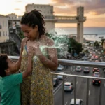 Fotografia horizontal de uma mãe parda com cabelos cacheados e seu filho de 4 anos em uma passarela em Salvador, Bahia, com o Elevador Lacerda ao fundo no pôr do sol. A criança toca o peito da mãe, de onde emana uma luz artística sutil em formato de rede celular, simbolizando o microquimerismo fetal e a regeneração de órgãos pelas células dos filhos. Cenário urbano brasileiro com prédios históricos em bokeh dourado, representando a conexão biológica eterna