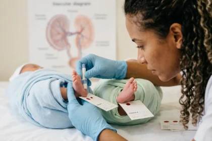 Close-up fotográfico e intimista focado nas mãos de uma enfermeira pediátrica (com luvas de látex azul claro) segurando, com delicadeza, dois pezinhos minúsculos e idênticos. Cada pezinho pertence a um bebê gêmeo diferente, cujas perninhas estão suavemente entrelaçadas em primeiro plano. A enfermeira está realizando a coleta em um dos pezinhos, e a outra mão segura o segundo, indicando que a coleta é dupla. Ao lado, vemos dois cartões de coleta separados (papel filtro), cada um com o nome 'Gêmeo 1' e 'Gêmeo 2'. No fundo, um borrão poético da placenta desfocada com o detalhe sutil da transfusão de sangue que eles dividem. A luz natural morna ilumina a cena, capturando a textura da pele e a conexão no olhar de ambos. O fundo é um borrão poético (bokeh) da casa